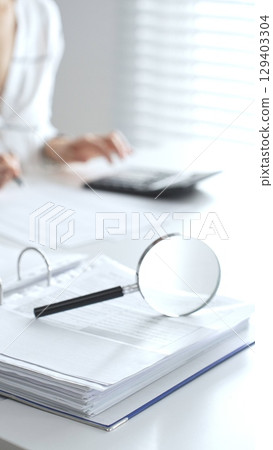 Magnifying glass on financial documents is in focus while an accountant using a calculator and taking notes, emphasizing accounting, finance, close up vertical view. Audit and taxes 129403304