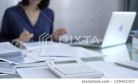 Magnifying glass, white calculator, pen and documents are lying on accountant's desk. Businesswoman wearing blue dress is working with documents on the background. Business and audit concept 129403307