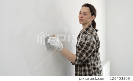 Woman wearing protective gloves and checkered shirt smoothing putty on white wall, spreading compound with spatula during home renovation, displaying practical home improvement skills 129403309