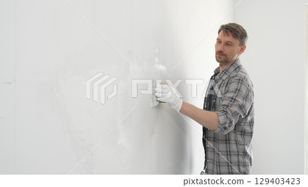 Construction worker spreading plaster on white wall, wearing protective gloves and t-shirt during home renovation project, working with professional skill and focus 129403423