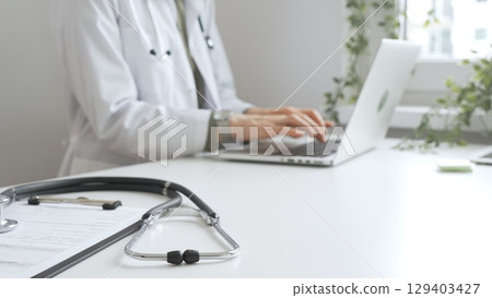 Stethoscope over a medical records are lying on the desk opposite the doctor is typing on a laptop with a in a medical office, suggesting online healthcare or telemedicine services. Medicine concept 129403427
