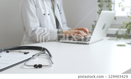 Stethoscope over a medical records are lying on the desk opposite the doctor is typing on a laptop with a in a medical office, suggesting online healthcare or telemedicine services. Medicine concept Stethoscope over a medical records are lying on the desk opposite the doctor is typing on a laptop with a in a medical office, suggesting online healthcare or telemedicine services. Medicine concept 129403429