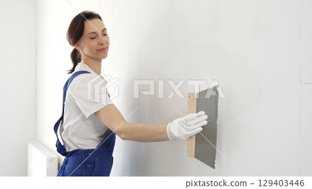 Woman construction worker wearing protective gloves and blue construction coveralls, spreading plaster smoothly across wall using professional drywall taping knife during renovation project. Portrait 129403446