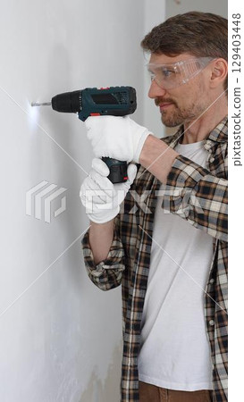 Middle aged male worker wearing checked shirt and white gloves, is drilling hole in a wall with cordless drill during home renovation work. Vertical portrait view Middle aged male worker wearing checked shirt and white gloves, is drilling hole in a wall with cordless drill during home renovation work. Vertical portrait view 129403448