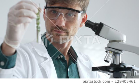 Adult male scientist in mask and gloves holding test tube with green plant sample, conducting laboratory research with a microscope. Medicine, healthcare and science concept Adult male scientist in mask and gloves holding test tube with green plant sample, conducting laboratory research with a microscope. Medicine, healthcare and science concept 129403461