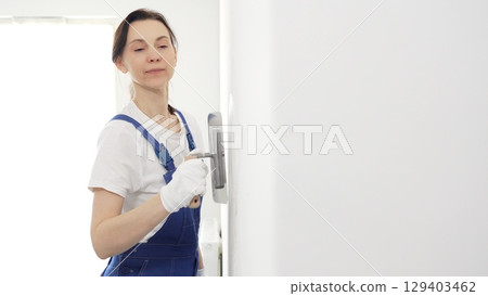 Professional female construction worker wearing blue coveralls is using a finishing trowel to carefully apply plaster on a wall, demonstrating expertise in home renovation and interior finishing 129403462