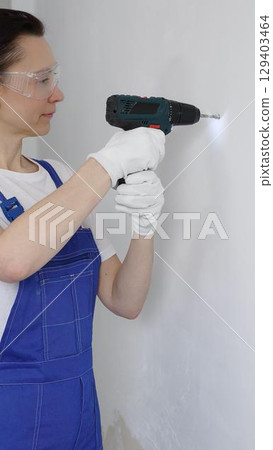 Professional female worker wearing blue overall, safety glasses and gloves, is using cordless drill to make hole in white wall during renovation work. Portrait view 129403464