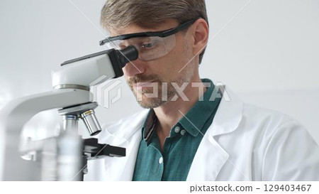 Adult male scientist wearing protective glasses and lab coat analyzing samples with a microscope in a modern laboratory setting. Medicine, healthcare and science concept 129403467