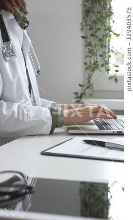 Doctor woman wearing white lab coat over green shirt, and stethoscope, is typing on laptop, documenting patient information. Medicine and health care concept 129403576