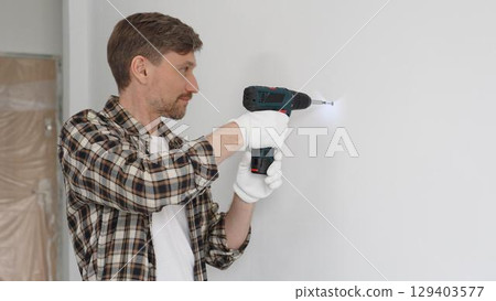 Middle aged man wearing checked shirt and white gloves, is drilling hole in a wall with cordless drill during home renovation work. Portrait view 129403577
