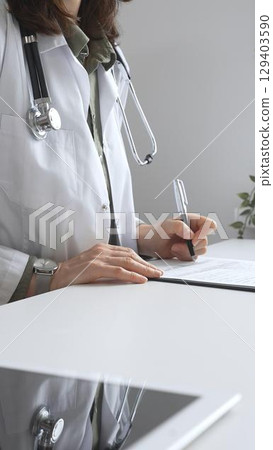 Professional female doctor wearing white lab coat and stethoscope documenting patient information on clipboard near laptop in clinic workspace. Medicine and health care concept Professional female doctor wearing white lab coat and stethoscope documenting patient information on clipboard near laptop in clinic workspace. Medicine and health care concept 129403590