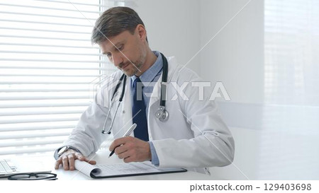 A male doctor in a lab coat with a stethoscope sits at a table in a bright medical office and uses a laptop while taking notes on a clipboard for patient care and treatment 129403698