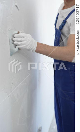 Close-up of man construction worker wearing gloves and blue overalls is using a putty knife to apply plaster on a white wall, performing home renovation and improvement 129403717
