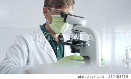 Adult male scientific professional analyzing sample through microscope, wearing green protective gear in sterile laboratory setting. Medicine, healthcare and science concept 129403719
