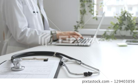 Stethoscope and medical records are on the desk opposite the doctor is typing on a laptop with a in a medical office, suggesting online healthcare or telemedicine services. Medicine concept 129403856
