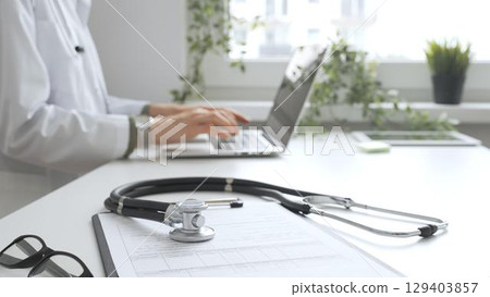Stethoscope over a medical records are lying on the desk opposite the doctor is typing on a laptop with a in a medical office, suggesting online healthcare or telemedicine services. Medicine concept 129403857