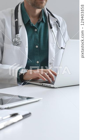 Doctor man wearing a white coat over green shirt, and stethoscope is typing on a laptop in medical office, showcasing the intersection of healthcare and technology, vertical close up view. Medicine 129403858