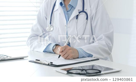 Female doctor documents patient information while working with digital devices at minimalist white workstation in medical office. Medicine and healthcare concept 129403861