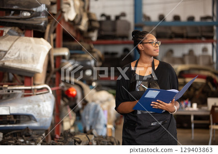 Confident african american female worker holding blue folder while inspecting car parts warehouse. Concept of automotive maintenance, quality control industrial service. 129403863