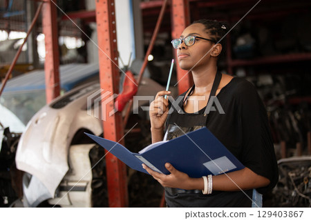 Confident african american female worker holding blue folder while inspecting car parts warehouse. Concept of automotive maintenance, quality control industrial service. Confident african american female worker holding blue folder while inspecting car parts warehouse. Concept of automotive maintenance, quality control industrial service. 129403867
