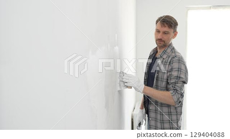 Construction worker wearing gloves and using a putty knife to plaster a white wall, performing home renovation and improvement tasks with precision 129404088