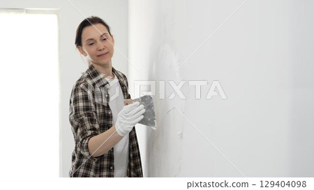 Woman wearing protective gloves and checkered shirt smoothing putty on white wall, spreading compound with spatula during home renovation, displaying practical home improvement skills Woman wearing protective gloves and checkered shirt smoothing putty on white wall, spreading compound with spatula during home renovation, displaying practical home improvement skills 129404098