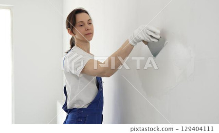 Female construction worker spreading finishing putty on white wall, wearing protective gloves and blue work overall while smoothing surface with putty knife for precise home repair 129404111