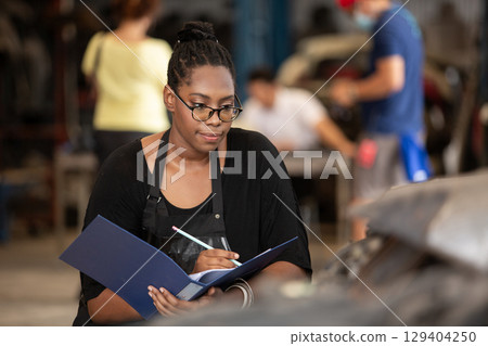 Confident african american female worker holding blue folder while inspecting car parts warehouse. Concept of automotive maintenance, quality control industrial service. Confident african american female worker holding blue folder while inspecting car parts warehouse. Concept of automotive maintenance, quality control industrial service. 129404250