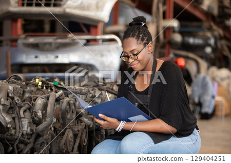 Confident african american female worker holding blue folder while inspecting car parts warehouse. Concept of automotive maintenance, quality control industrial service. 129404251