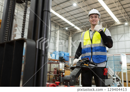 Confident warehouse worker operating forklift in industrial factory. Wearing safety white helmet and vest, he gives thumbs up showing job satisfaction and commitment to workplace safety. 129404413