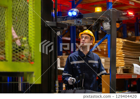 Industrial worker operating forklift in warehouse while wearing safety gear and uniform. Focused male employee managing inventory logistics and lifting materials in storage facility. Industrial worker operating forklift in warehouse while wearing safety gear and uniform. Focused male employee managing inventory logistics and lifting materials in storage facility. 129404420