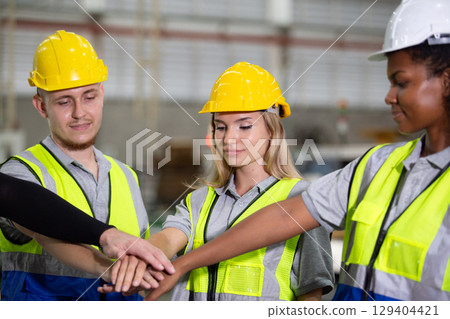Diverse industrial workers in safety uniforms and helmets stacking hands in teamwork gesture, showing unity, collaboration, and motivation at workplace in manufacturing or factory environment. 129404421