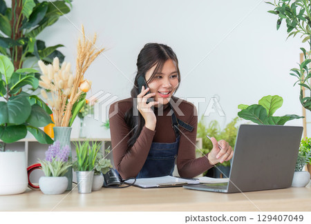 Customer Service. Young woman on the phone while working on a laptop in a plant-filled office. Customer Service. Young woman on the phone while working on a laptop in a plant-filled office. 129407049