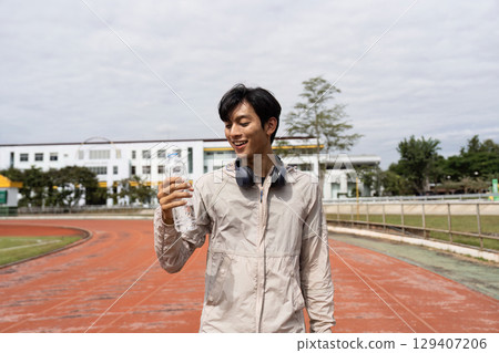Young Man Enjoying a Refreshing Break During a Jog on a Track, Embracing Wellness and Fitness with a Smile and a Bottle of Water 129407206