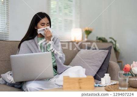 Young Woman Feeling Unwell at Home, Wrapped in Blanket, Using Laptop, and Blowing Nose with Tissue Young Woman Feeling Unwell at Home, Wrapped in Blanket, Using Laptop, and Blowing Nose with Tissue 129407224