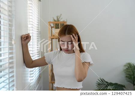 Young Woman Feeling Unwell at Home Leaning Against Wall in Bright Room with Plants and Shelves Young Woman Feeling Unwell at Home Leaning Against Wall in Bright Room with Plants and Shelves 129407425