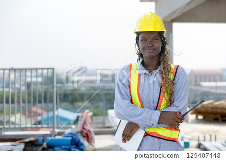 Portrait of female construction engineer standing confidently at outdoor site. Wearing safety gear and holding walkie-talkie and tablet, representing leadership, skill and professionalism. Portrait of female construction engineer standing confidently at outdoor site. Wearing safety gear and holding walkie-talkie and tablet, representing leadership, skill and professionalism. 129407448
