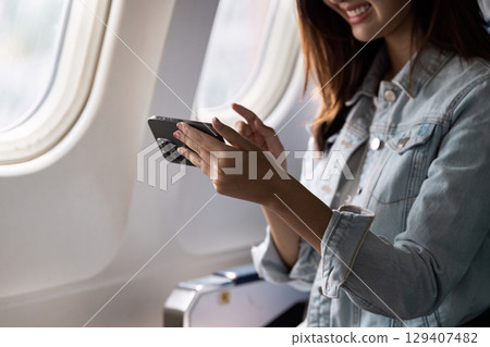 Woman Enjoying In-Flight Entertainment on Smartphone While Traveling on Airplane with Sunlight Streaming Through Window Woman Enjoying In-Flight Entertainment on Smartphone While Traveling on Airplane with Sunlight Streaming Through Window 129407482