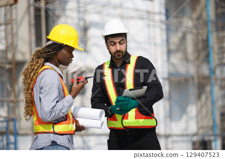 Two construction engineers working on-site, reviewing project data on tablet while discussing through walkie-talkie. Safety, planning and teamwork concept in outdoor construction environment. 129407523