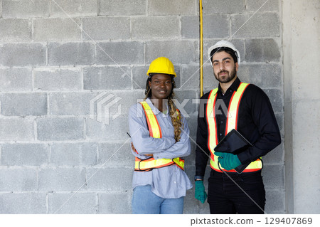 Portrait of two confident construction engineers standing at worksite. Wearing safety gear and holding a tablet, they represent leadership, teamwork, and professionalism in construction. 129407869