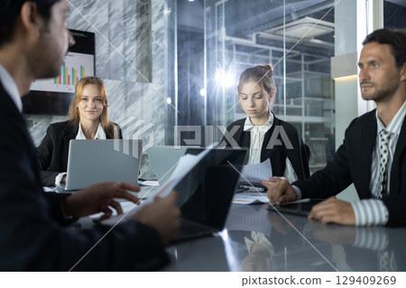 Business team in modern glass office engaged in meeting, reviewing documents and laptops, discussing data and strategies for corporate growth and professional collaboration. Business team in modern glass office engaged in meeting, reviewing documents and laptops, discussing data and strategies for corporate growth and professional collaboration. 129409269