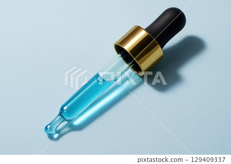 A clear glass dropper with blue liquid, black bulb, and gold collar on a light blue surface. 129409337