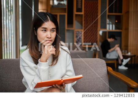 Young woman in a modern cafe, holding a notebook and pen, deep in thought. Warm, inviting atmosphere with wooden decor. 129412833