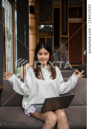 A young woman practices meditation with a laptop in a cozy living room, blending relaxation and technology. A young woman practices meditation with a laptop in a cozy living room, blending relaxation and technology. 129412842