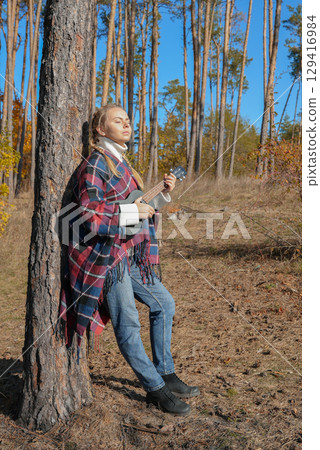 Portrait of smiling caucasian girl plays ukulele in autumnal park. Young woman plays guitar musical instrument outside in nature fall time. Audio music healing 129416984