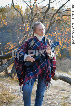 Portrait of smiling caucasian girl plays ukulele in autumnal park. Young woman plays guitar musical instrument outside in nature fall time. Audio music healing 129416986
