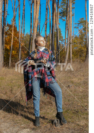 Blonde woman playing guitar ukulele outdoors in autumn forest. Concept of sound therapy, mental health and wellness rituals. Calmness tranquility audio-sensory practices. Aura farming 129416987