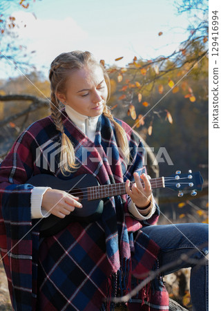 Beautiful blonde girl playing guitar ukulele and singing relaxing enjoying outdoors on sunny autumn day. Hippie woman in boho style training playing musical instrument. Fall leaves Beautiful blonde girl playing guitar ukulele and singing relaxing enjoying outdoors on sunny autumn day. Hippie woman in boho style training playing musical instrument. Fall leaves 129416994