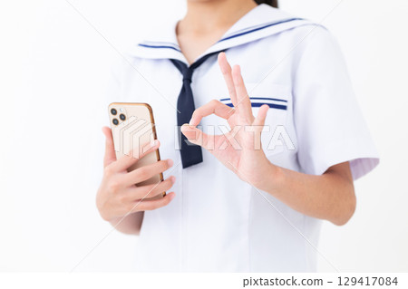 A junior high school girl in a summer school uniform holding a smartphone and making a circle sign with her fingers 129417084