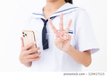 A junior high school girl in a summer school uniform holding a smartphone and pointing three fingers up 129417123